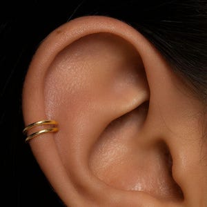May include: A close-up of a gold-coloured double ear cuff. The ear cuff is worn on the upper ear and has a smooth, polished finish. The background is a solid black.