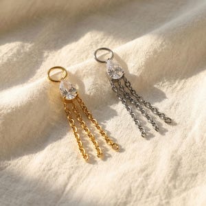 May include: Two earrings, one gold and one silver, each featuring a teardrop-shaped clear stone and three dangling chains. The earrings are set against a soft, off-white fabric background.