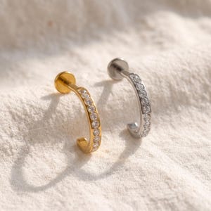 May include: Two curved labret studs featuring a row of clear, round-cut stones set along the front. One stud is finished in a polished gold tone, while the other is finished in a polished silver tone, each with a flat circular back disc.