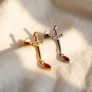 Peut inclure: Deux piercings labret en forme d'épée, l'un doré et l'autre argenté. Chaque bijou présente une petite pierre rouge ronde sur la garde et une pierre rouge taille marquise suspendue à la pointe, fixée sur une tige à embout plat.