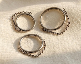 May include: Three silver rings with attached chains. The rings are circular and have a simple, minimalist design. The chains are delicate and connect to the rings. The rings are displayed on a cream-coloured fabric.