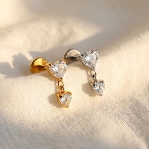 May include: Two heart-shaped earrings. One earring is gold-toned, with a small heart-shaped gem dangling below a larger heart-shaped gem. The other earring is silver-toned, with two heart-shaped gems. Both earrings have a post backing.