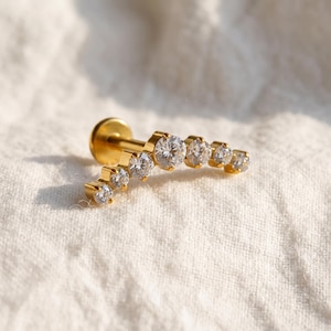 May include: A gold-toned labret stud earring featuring a curved bar design set with seven clear, round-cut cubic zirconia stones that graduate in size, attached to a flat circular backing post for secure placement in a piercing.