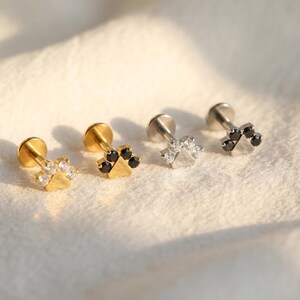 May include: Four small stud earrings with paw print designs. Two are gold-toned with clear and black stones, and two are silver-toned with clear and black stones. The earrings are displayed on a soft, white surface.