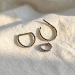 May include: Three silver-colored, D-shaped hoop earrings of varying sizes are displayed on a white, textured surface. The earrings have a simple, minimalist design, with a straight bar across the top of the D.