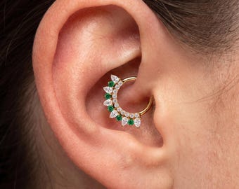 May include: A gold hoop earring featuring a semi-circular arrangement of alternating pear-cut white stones and round-cut green stones. The jewellery is positioned in the conch area of an ear, showcasing a curved metal band with a decorative cluster.