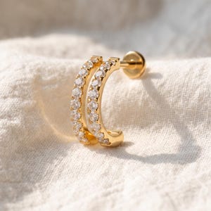 Peut inclure: Une boucle d'oreille créole dorée ornée de deux rangées parallèles de pierres claires taillées en rond. Le bijou présente une forme semi-circulaire incurvée avec une tige plate et un fermoir rond, affichant une finition métallique polie.