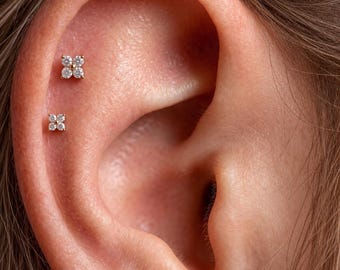 May include: Two gold-toned stud earrings, each featuring a four-petaled flower design composed of four small, clear, round-cut stones. The earrings are positioned vertically along the upper cartilage of the ear, showcasing a delicate, symmetrical look.