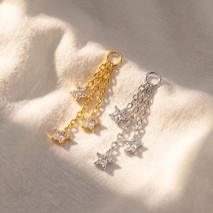 May include: Two star-shaped charm pendants, one gold-toned and one silver-toned, each with three dangling chains. Each chain has a star-shaped, clear crystal. The pendants are on a white, textured surface.