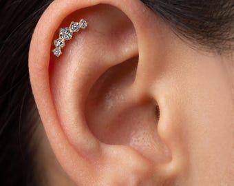 May include: A gold-toned ear cartilage stud featuring a curved row of five clear, round-cut stones of varying sizes. The jewellery is positioned along the upper edge of the ear, with the largest stone centred among four smaller stones.
