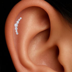 May include: A curved ear piercing with seven clear, round gemstones. The gemstones are arranged in descending size, creating a sparkling design. The piercing is set against the backdrop of a person's ear.