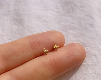 May include: Two small, gold-coloured star-shaped stud earrings. The earrings are displayed on a finger against a soft, light-coloured background. The earrings are a simple, minimalist design.