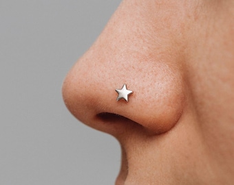 May include: A small, polished silver-toned star-shaped nose stud. The jewelry features a five-pointed star design with a smooth, reflective metallic surface, positioned securely on the side of the nostril for a simple and clean appearance.