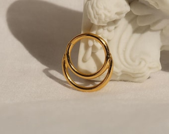 May include: A gold-coloured, double-hoop earring. The earring is circular with a hinged clasp. The jewellery is displayed on a white surface with a white sculpture in the background. The earring is made of a shiny, reflective metal.