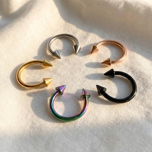 May include: A selection of horseshoe-shaped body jewellery with conical spikes. The jewellery is arranged in a circle. Colours include silver, gold, rose gold, black, and iridescent. The spikes are pointed and symmetrical, and the jewellery is made of metal.
