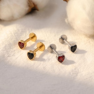 May include: Four small stud earrings with heart-shaped gemstones. Two earrings have gold-colored posts and settings, one with a red gemstone and one with a black gemstone. The other two earrings have silver-colored posts and settings, one with a red gemstone and one with a black gemstone.