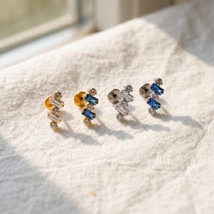 May include: Four stud earrings featuring a vertical arrangement of two baguette-cut stones and two round-cut stones. Two earrings are gold-toned with clear stones, and two are silver-toned with blue stones, all with flat back posts.