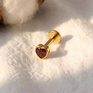 May include: A gold-colored lip piercing stud with a heart-shaped, deep red gemstone. The stud has a flat back and a post for insertion. The heart-shaped gem is set in a gold bezel. The jewelry is displayed on a white, textured surface.