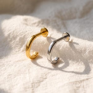 Könnte beinhalten: Zwei Labret-Piercings, eines in poliertem Goldton und eines in poliertem Silberton. Jedes Stück verfügt über einen glatten, gebogenen Stab mit einer flachen, kreisförmigen Scheibenbasis an einem Ende und einer abgerundeten, halbkreisförmigen Hakenform am anderen Ende.