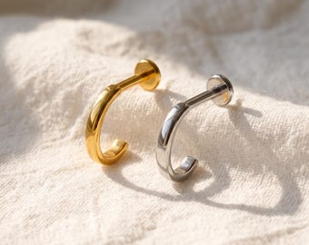 May include: Two labret piercings, one in a polished gold tone and one in a polished silver tone. Each piece features a smooth, curved post with a flat circular disc base at one end and a rounded, semi-circular hook shape at the other end.
