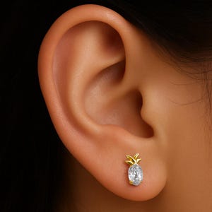 May include: A gold-colored pineapple stud earring with a clear, oval-shaped gemstone. The earring is worn on the ear. The pineapple design features a textured top and a smooth, polished body.
