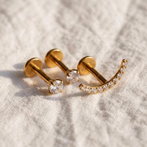 May include: Three gold-toned flat back labret studs. Two studs feature a single round clear gemstone, while the third stud has a curved bar design set with a row of small clear gemstones. The metal posts are straight with circular backings.