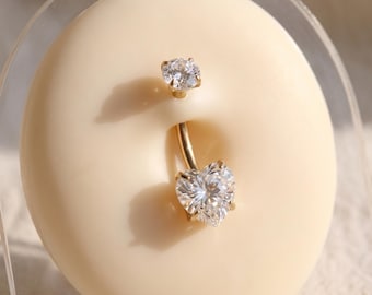 May include: A gold navel piercing ring featuring a small round clear gemstone at the top and a larger heart-shaped clear gemstone at the bottom. The jewellery is set in a smooth, light-toned circular display form, showcasing the curved metal bar.