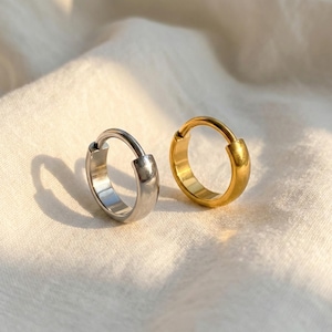 May include: Two hoop earrings, one silver and one gold, are shown on a white, textured surface. The earrings have a simple, rounded design, with a hinged closure. The gold earring has a shiny finish, while the silver earring has a matte finish.