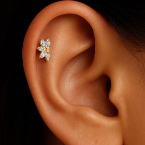May include: A gold-colored ear piercing stud in the shape of a flower, with six clear, marquise-cut gemstones. The stud is set against the skin of an ear, highlighting the jewelry's design and placement.