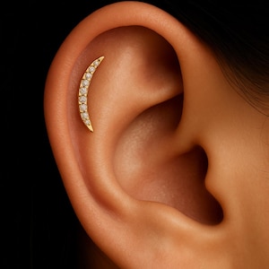 Può includere: Un orecchino curvo color oro con una fila di piccole pietre preziose trasparenti. L'orecchino ha la forma di una mezzaluna ed è inserito nell'orecchio di una persona. Lo sfondo è un primo piano dell'orecchio e della pelle circostante.