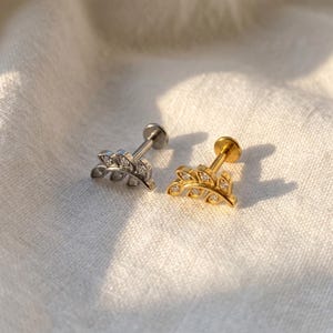 Op de afbeelding: Twee oorbellen in de vorm van een blad. De ene is zilverkleurig, de andere goudkleurig. Beide hebben kleine, heldere edelstenen. De oorbellen hebben een platte achterkant en een pin voor piercing.