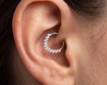 May include: A silver-toned circular hoop earring featuring a row of small, clear, pear-shaped stones along one side. The jewellery is positioned within the inner cartilage of the ear, specifically designed as a daith or septum piercing ring.