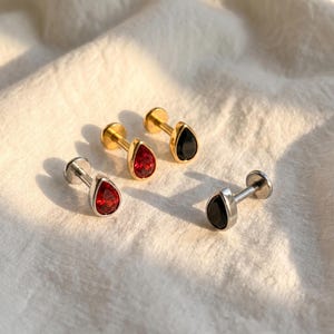 May include: Four stud earrings featuring teardrop-shaped gemstones. Two earrings have gold-toned posts with one red and one black stone. Two earrings have silver-toned posts with one red and one black stone. Each earring has a flat circular back.