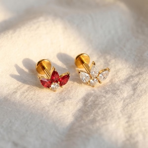 May include: Two gold-coloured stud earrings. One earring features a cluster of red gemstones and a central clear gemstone. The other earring has a cluster of clear gemstones. Both earrings are on a white, textured surface.