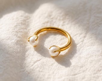 May include: A gold-toned open ring featuring two white spherical pearls, one at each end of the band. The pearls are held in place by small metal prongs, creating a circular design with a gap between the two pearl accents on the band.