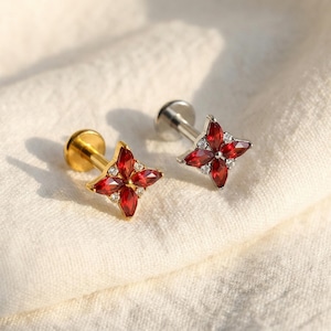 Puede incluir: Dos pendientes con diseño de flor de cuatro pétalos. Uno tiene un poste y engaste dorado, el otro plateado. Ambos presentan gemas rojas vibrantes y pequeños acentos blancos.