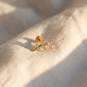 Könnte beinhalten: Ein goldfarbener Ohrring mit Blattdesign. Der Ohrring hat einen kleinen Stecker und einen gebogenen Zweig mit mehreren klaren, funkelnden Steinen. Das Blattdesign ist detailliert und filigran, vor einem weichen, cremefarbenen Hintergrund.
