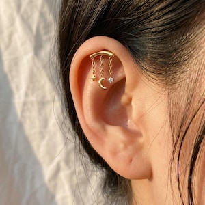 May include: Gold-coloured ear jewellery featuring a curved bar with three dangling charms: a star, a crescent moon, and a small, round, clear gem. The jewellery is worn on the upper ear cartilage.