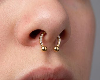 May include: A gold horseshoe-shaped septum ring featuring a row of small, clear, sparkling stones embedded along the curved band, finished with polished gold spheres at both ends, positioned securely within the nasal septum.