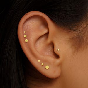 Peut inclure: Gros plan sur une oreille ornée de plusieurs petits clous d'oreilles dorés. Les boucles d'oreilles sont de différentes tailles, créant un effet superposé. L'oreille est mise en valeur sur un fond sombre.