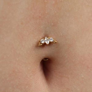 May include: A gold navel piercing jewelry piece featuring a central marquise-cut clear gemstone flanked by two smaller round clear gemstones, accented with tiny gold spheres on each side, positioned above the navel opening on skin.