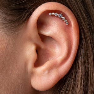 Puede incluir: Un pendiente de cartílago para la oreja con forma curva, que presenta una hilera de siete piedras preciosas redondas y transparentes engastadas en metal. La joya sigue el contorno natural del cartílago superior de la oreja.