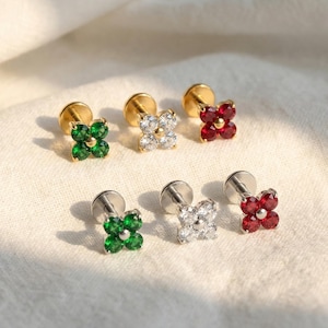 May include: A collection of stud earrings featuring four-leaf clover designs. The earrings are available in gold and silver tones, with green, clear, and red gemstone accents. The earrings are displayed on a white surface.