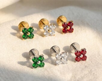 May include: Stud earrings with four-leaf clover designs in gold and silver. The earrings have green, clear, and red gemstone details. The earrings are presented on a white surface.
