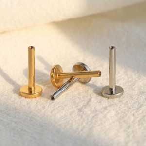 Può includere: Sono mostrati tre piercing per labbra. Due sono color oro e uno color argento. Ogni piercing ha una base piatta e rotonda e un perno cilindrico con un'estremità filettata. I piercing sono esposti su una superficie bianca e strutturata.