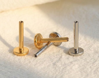 May include: Three lip piercing studs are shown. Two are gold-coloured, and one is silver-coloured. Each stud has a flat, round base and a cylindrical post with a threaded end. The studs are displayed on a white, textured surface.