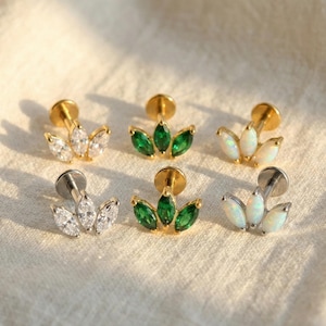 Puede incluir: Una variedad de pendientes de botón con un diseño de tres pétalos. Los pendientes tienen postes dorados y plateados, y presentan piedras de talla marquesa transparentes, verdes y opalescentes. Se exhiben sobre un fondo de tela neutra.