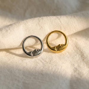 May include: Two crescent moon-shaped rings, one silver and one gold, are displayed on a white fabric. Each ring features a detailed face design. The rings are circular and appear to be made of metal.