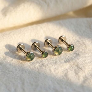 Op de afbeelding: Vier zilverkleurige, platte oorbellen met ronde, groene edelsteen centra. De oorbellen zijn in een rij gerangschikt, elk met een staafje en een kleine, platte schijf aan de achterkant. De edelstenen hebben een mosachtig, natuurlijk uiterlijk.
