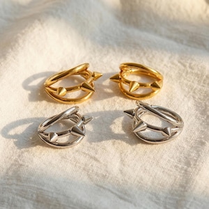 May include: Four rings, two gold and two silver, each with a double band and spike and stud details. The rings are displayed on a white, textured surface, with soft lighting.